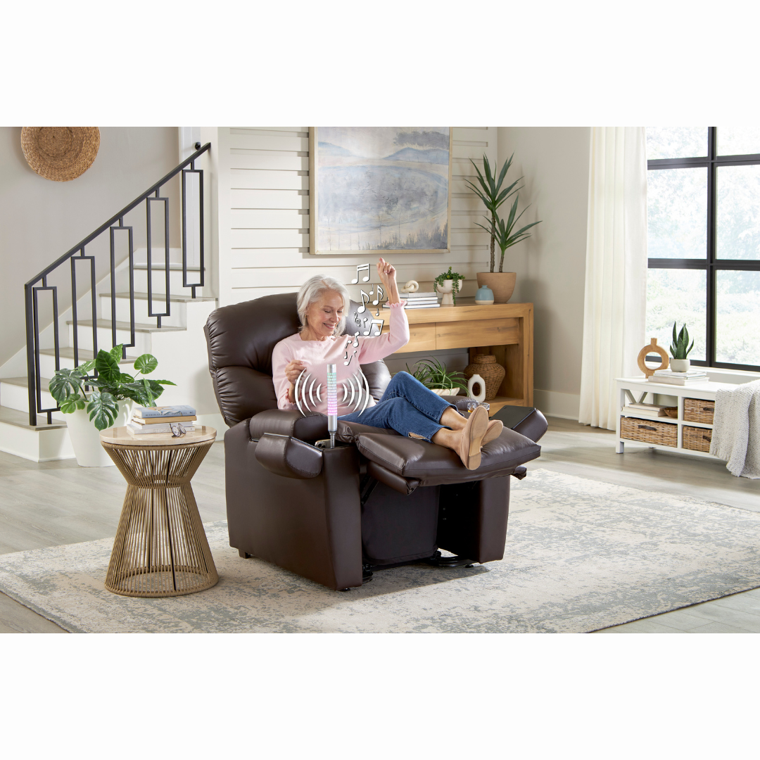 An elderly woman relaxes in a Golden Cloud+ PR511 Recliner with Lift Assist - Medium Wide, smiling as she listens to music with headphones. Her legs are crossed, enjoying the 4-Zone Comfort and AutoDrive hand control in her bright, plant-adorned living room while musical notes float around.