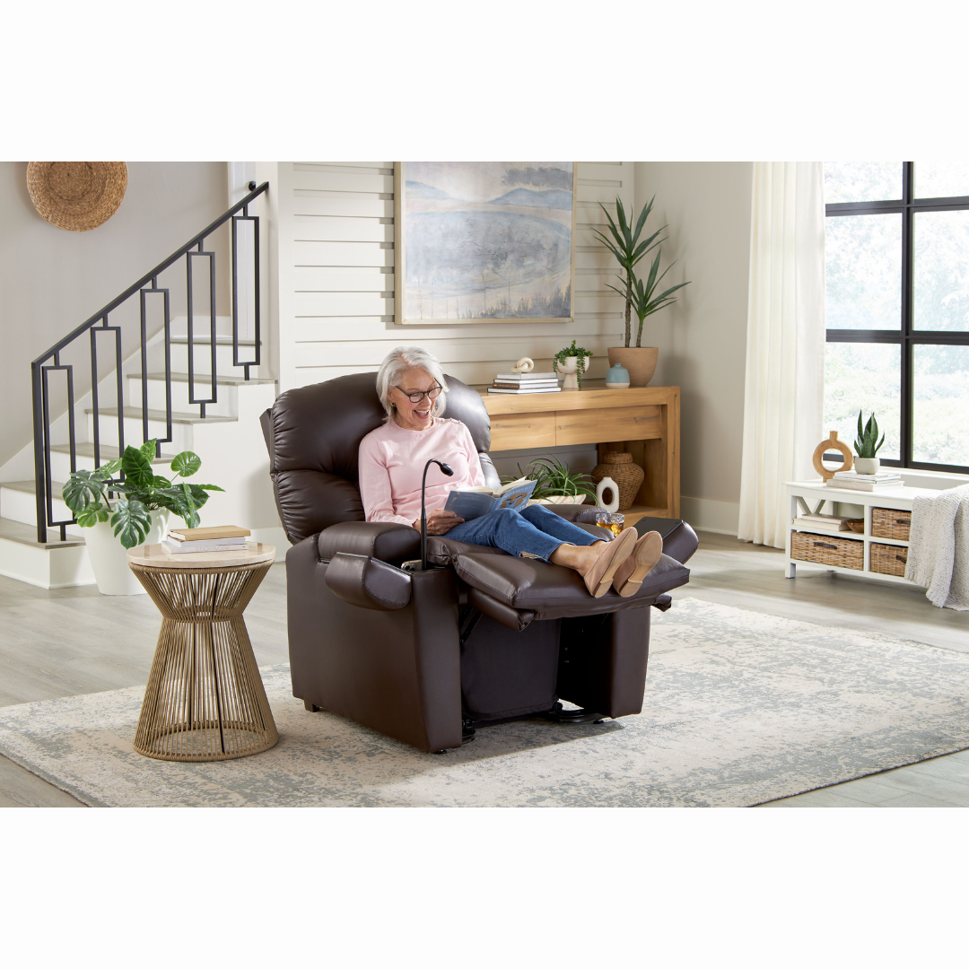 A gray-haired person with glasses relaxes in a Golden Cloud+ PR511 Recliner with Lift Assist - Medium Wide, reading in a cozy living room. The room includes a staircase, potted plants, a wooden side table, and a large window letting light stream in.