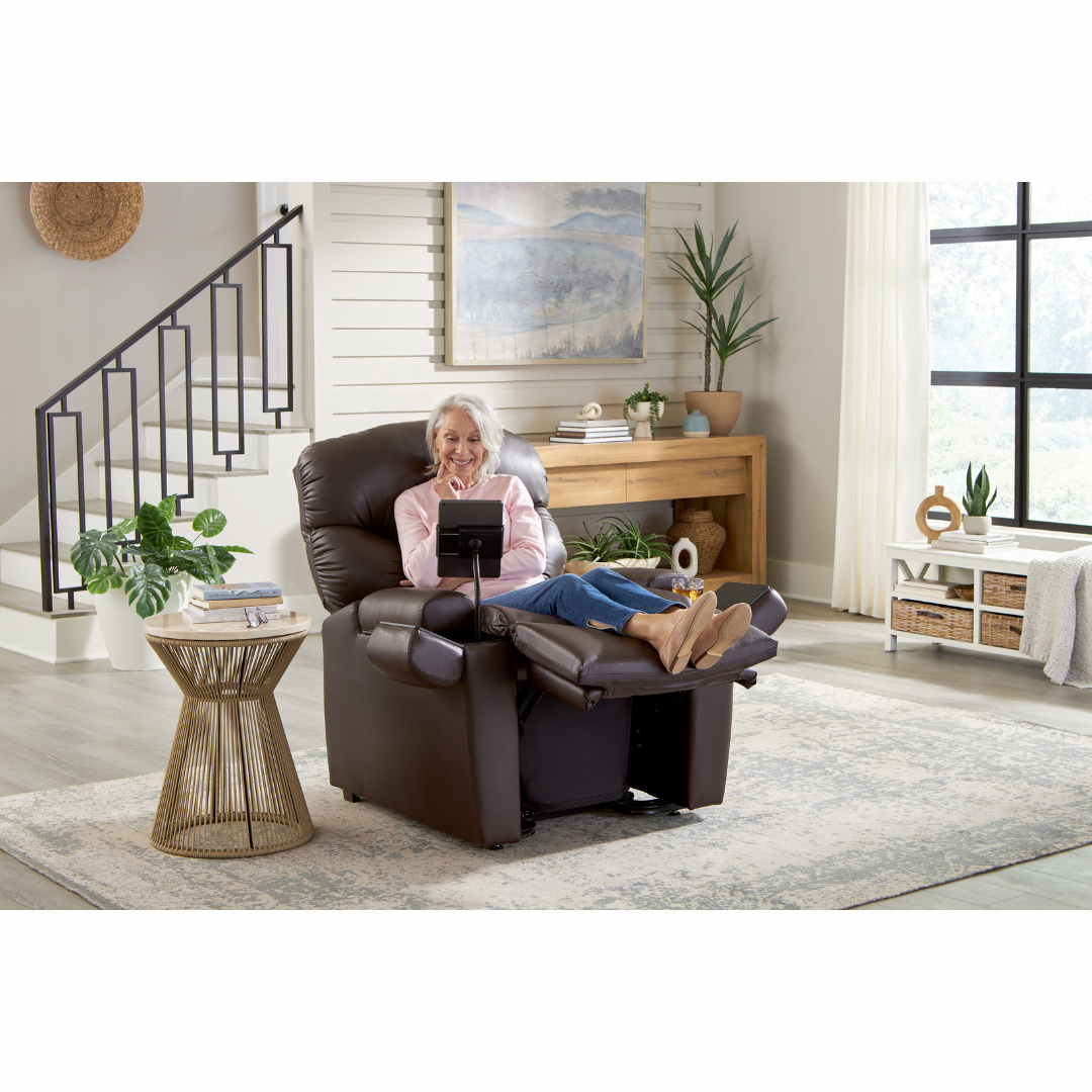A person with gray hair sits in a dark brown Golden Cloud+ PR511 Recliner with Lift Assist - Medium Wide, holding a tablet. The room includes a light rug, a wooden side table with a plant, and stairs in the background. Light pours through large windows framed by white curtains.