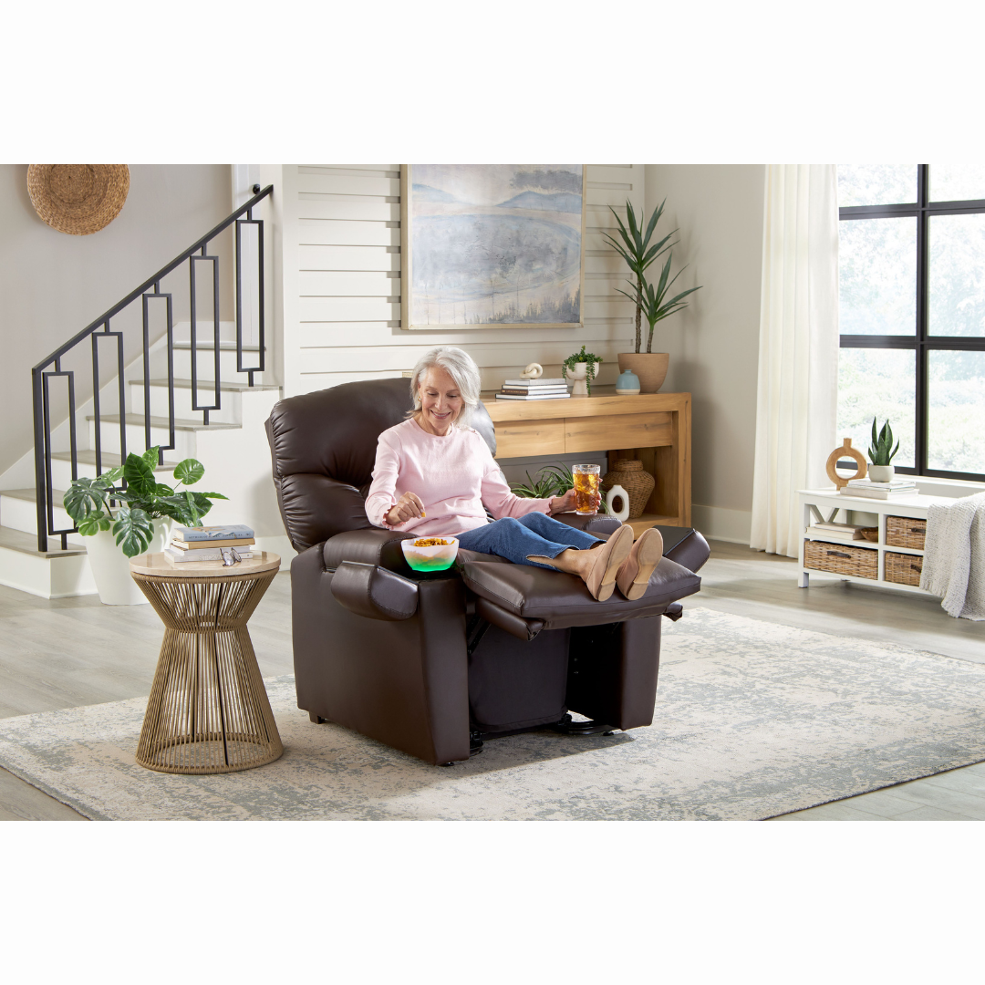 An elderly woman relaxes in a Golden Cloud+ PR511 Recliner with Lift Assist - Medium Wide, enjoying snacks from a cup on her lap. Her smile reflects the cozy living rooms warmth, featuring modern decor, plants, and a round side table by the large window. The 4-Zone Comfort Zone feature enhances her serene moment.