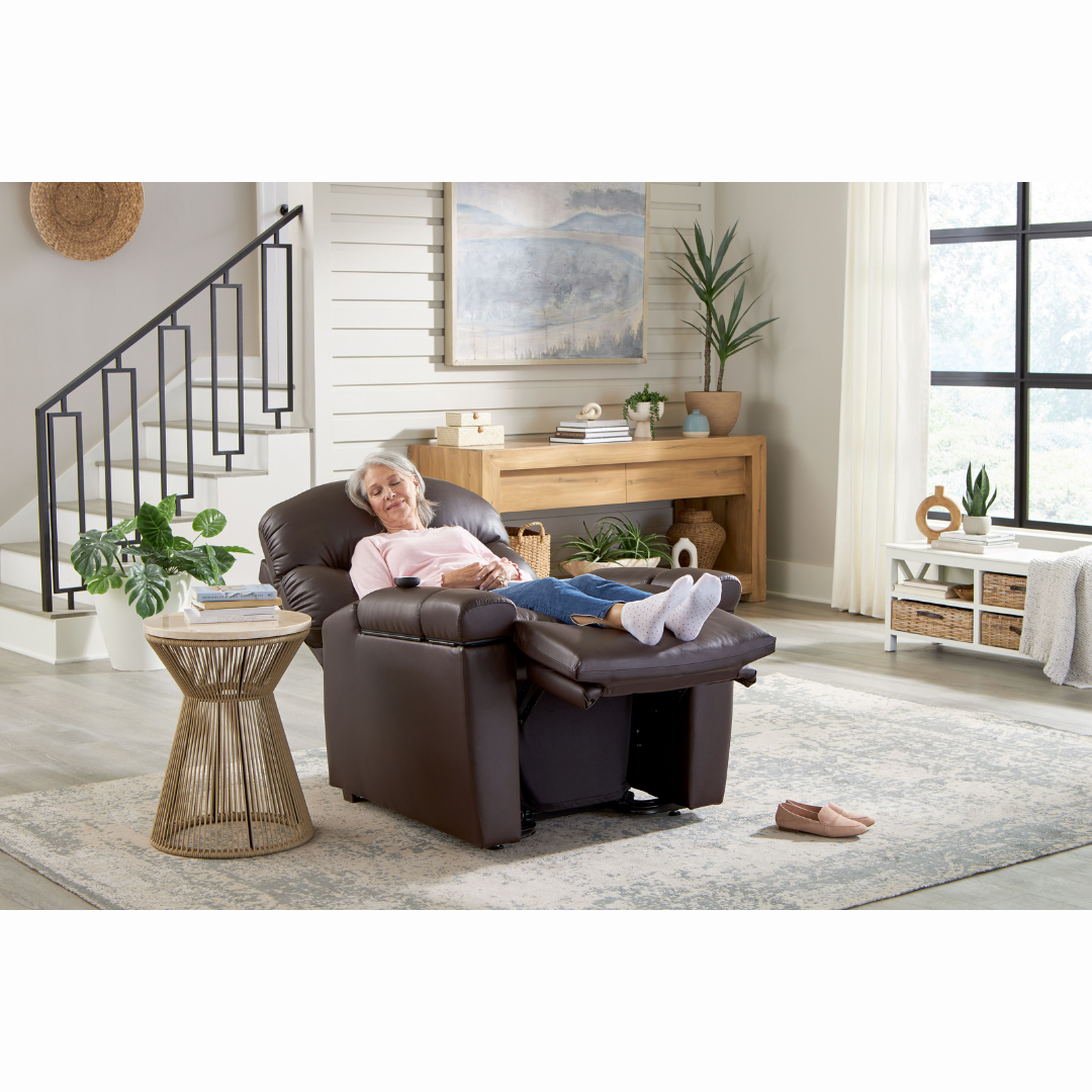 An elderly woman relaxes in a Golden Cloud+ PR511 Recliner with Lift Assist - Medium Wide, reading a book in a cozy, modern living room. Potted plants adorn the space as natural light floods through the window. A side table holds a cup, and slippers rest on the nearby rug.
