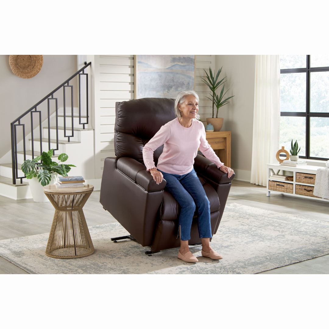 An elderly person in a pink sweater and jeans smiles warmly as they are gently assisted to stand from a Golden Cloud+ PR511 Recliner with Lift Assist - Medium Wide using the intuitive AutoDrive hand control. The bright living room features modern decor with a woven side table and stairway in the background.
