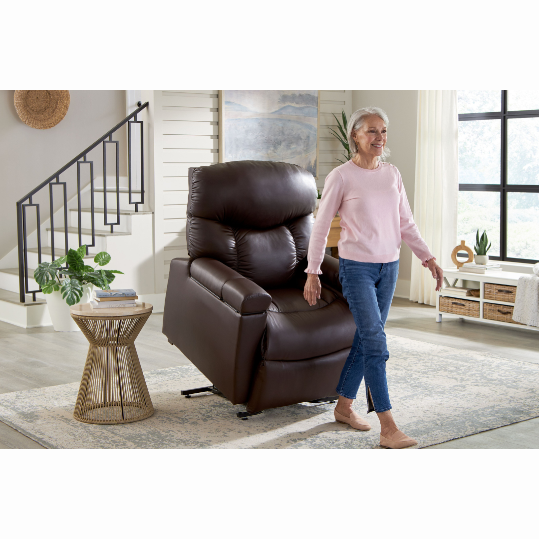 An elderly woman in a pink sweater and jeans stands beside her Golden Cloud+ PR511 Recliner with Lift Assist - Medium Wide, using the AutoDrive hand control in a bright, modern living room. A woven side table surrounded by plants bathes in natural light from a large window, enhancing the 4-Zone Comfort Zone.