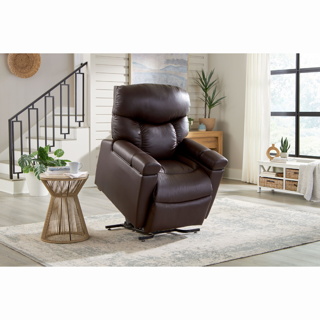 A cozy living room showcases the Golden Cloud+ PR511 Recliner with Lift Assist (Medium Wide) centered on a light rug. A nearby round wooden table holds a vase of greenery. Large windows and a black-railed staircase enhance brightness and depth in the space.