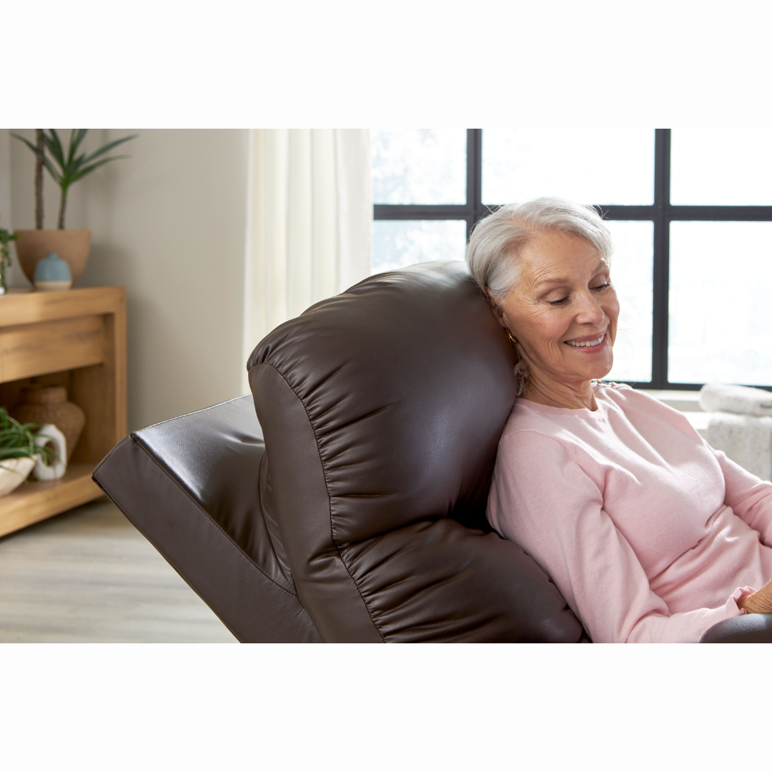 An elderly woman with gray hair relaxes contentedly in her pink sweater on a Golden Cloud+ PR511 Recliner with Lift Assist - Medium Wide. Indoors, a wooden shelf and large window let in soft natural light as she enjoys her 4-Zone Comfort Zone experience.