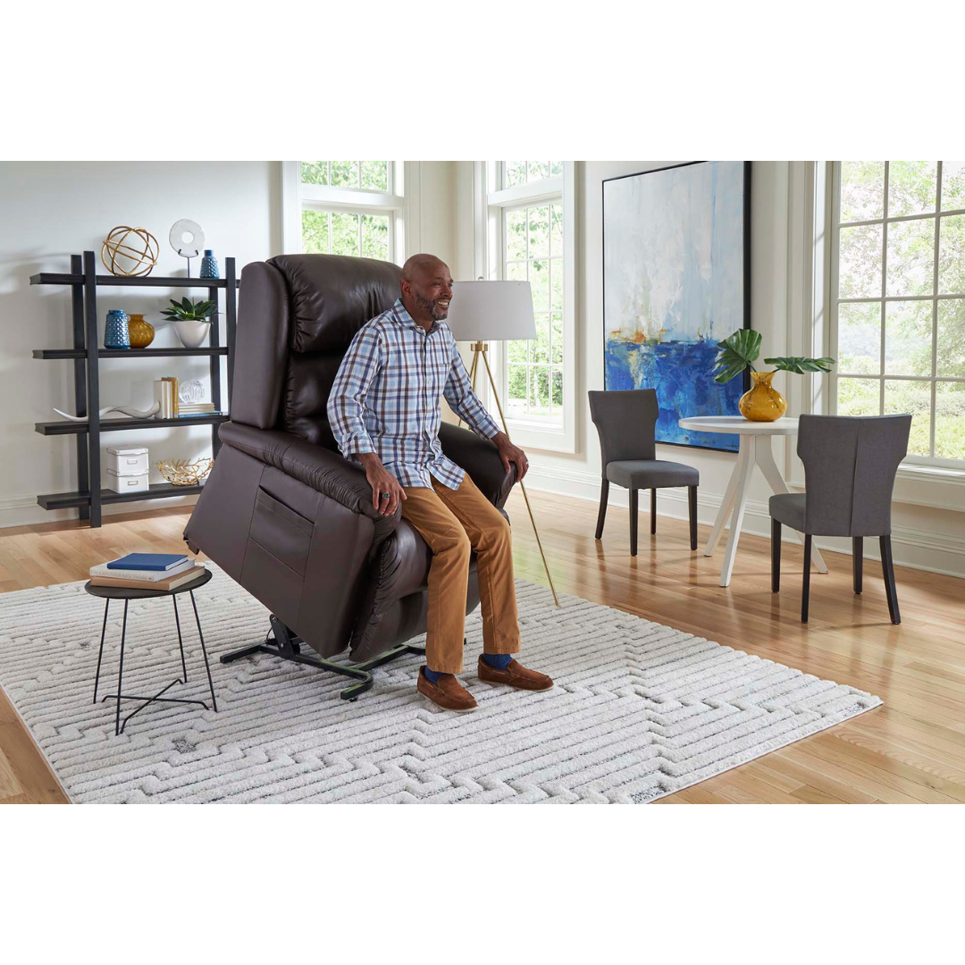 Someone relaxes in the Golden Tech PR766 Relaxer MaxiComfort Recliner With Lift Assist - Small in a bright living room with large windows. The space features a patterned rug, small round table, dining set, modern art, and a black shelf displaying decor for an ultimate relaxation experience.