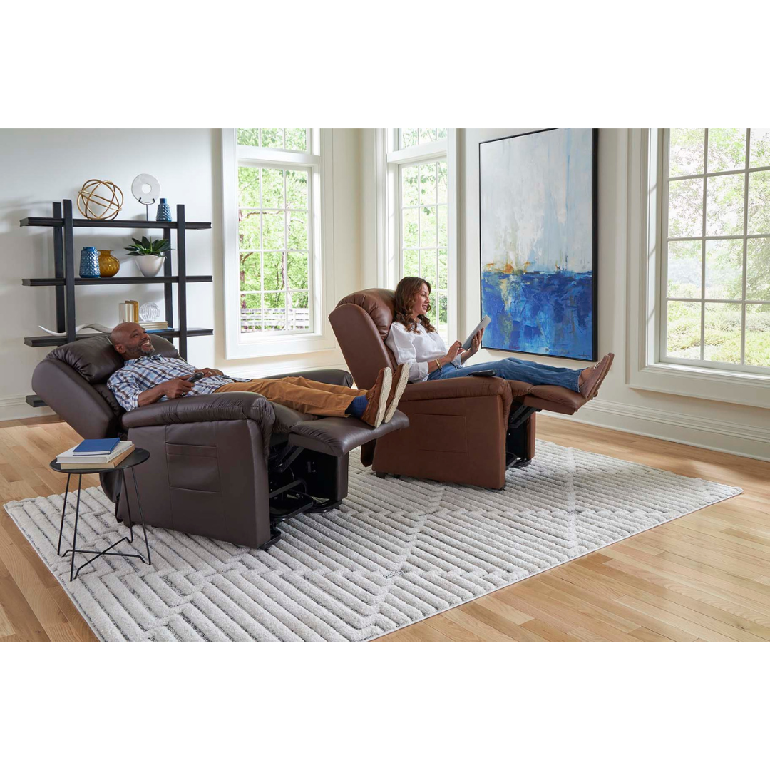 In a well-lit room with large windows, a man reclines peacefully with his eyes closed in his Golden Tech PR766 Relaxer MaxiComfort Recliner With Lift Assist - Small, using the programmable AutoDrive hand control, while a woman reads. A large abstract painting and shelf are in the background.