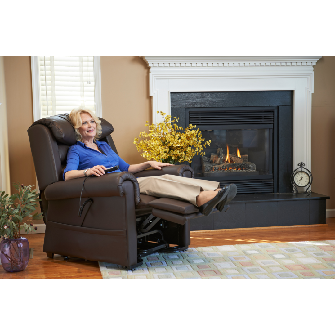 A person relaxes in a Golden Tech PR766 Relaxer MaxiComfort Recliner With Lift Assist - Small near a glowing fireplace. Light streams through the window, highlighting a decorative plant and clock, as they adjust the chairs settings with the Programmable AutoDrive Hand Control.