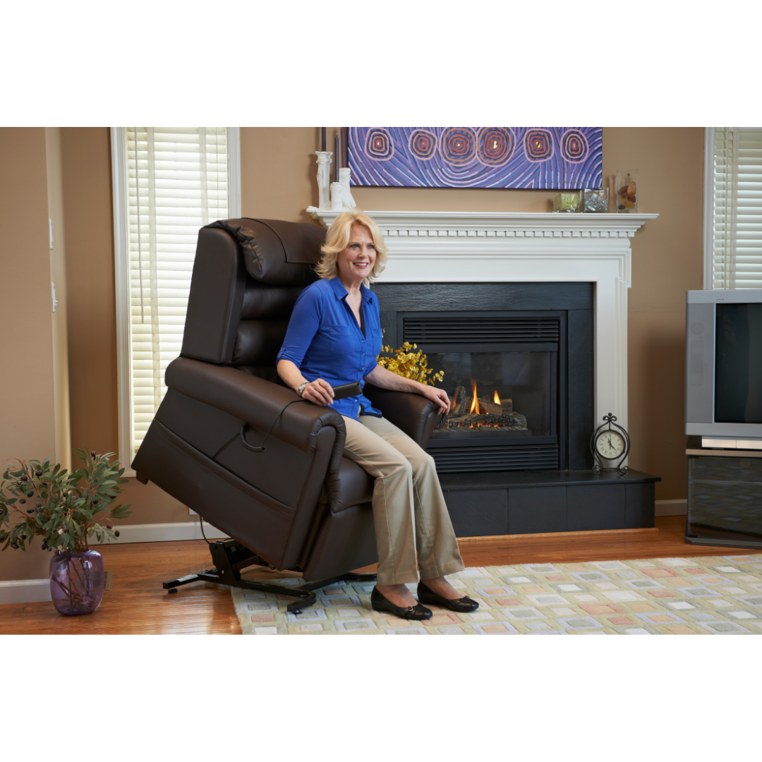 A woman in a blue blouse and beige pants sits on a Golden Tech PR766 Relaxer MaxiComfort Recliner With Lift Assist - Small. The living room has a lit fireplace with a colorful painting above it, a TV on the right, and a plant on the left. Shes smiling at the camera, enjoying her MaxiComfort Positioning.