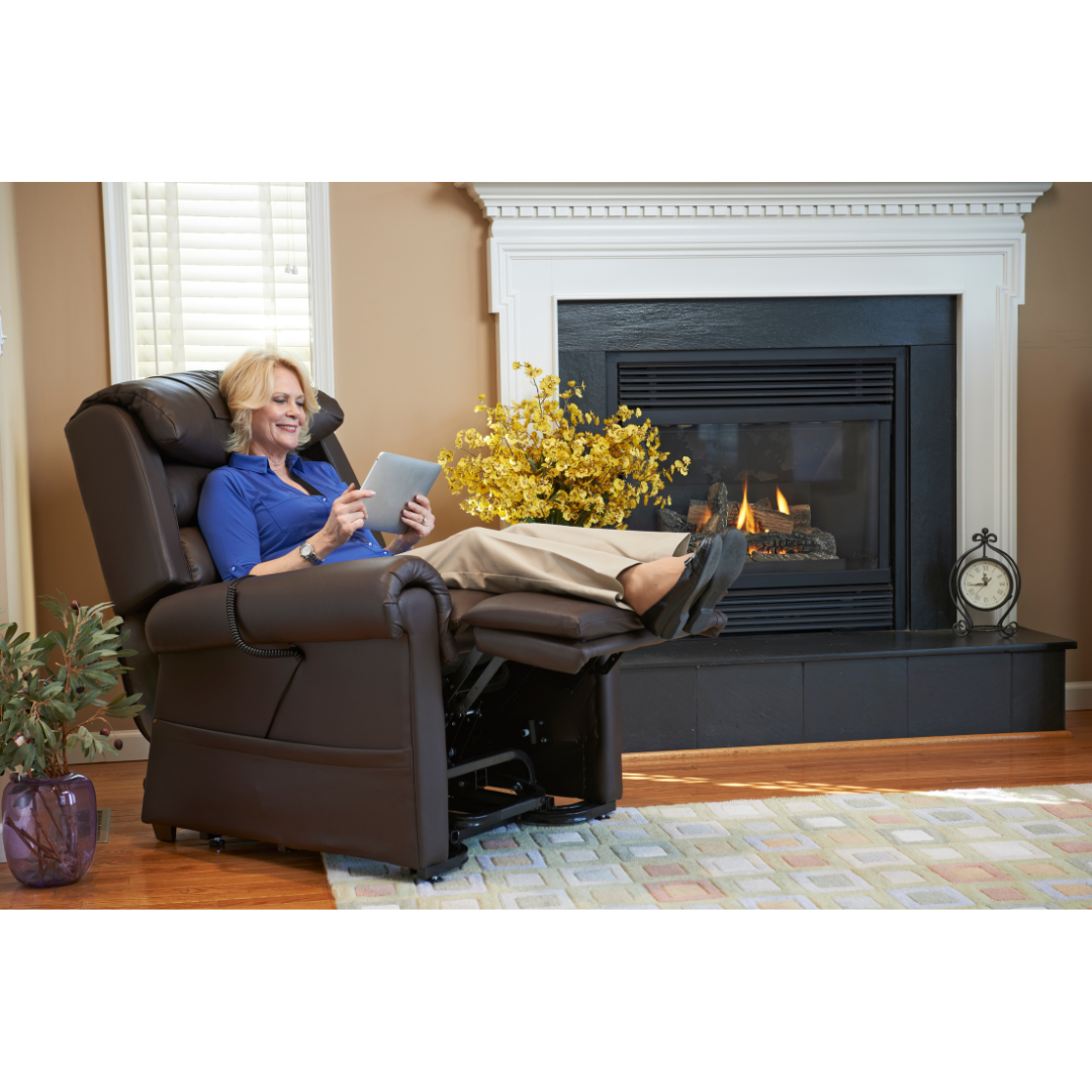 Sitting in a Golden Tech PR766 Relaxer MaxiComfort Recliner with Lift Assist - Small near a lit fireplace, they hold a tablet and smile. A plant, small clock, and patterned rug adorn the room, while MaxiComfort Positioning offers unmatched relaxation by the cozy fires warmth.