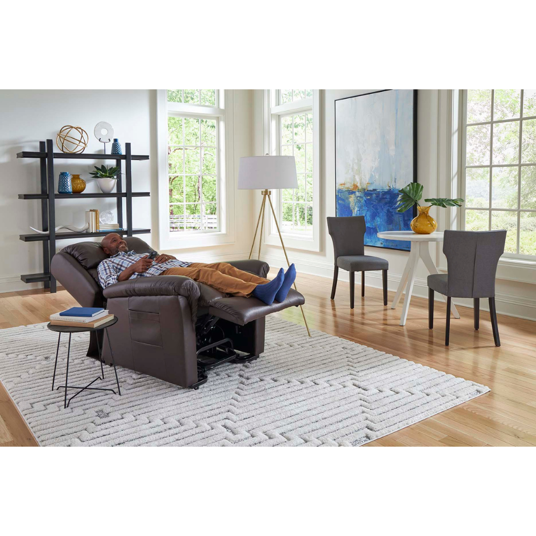 Relaxing in a Golden Tech PR766 Relaxer MaxiComfort Recliner With Lift Assist - Small, a person enjoys natural light from large windows. The modern living room boasts a patterned rug on wooden floors, an abstract painting, and features a small table with lamp near an elegant dining set.