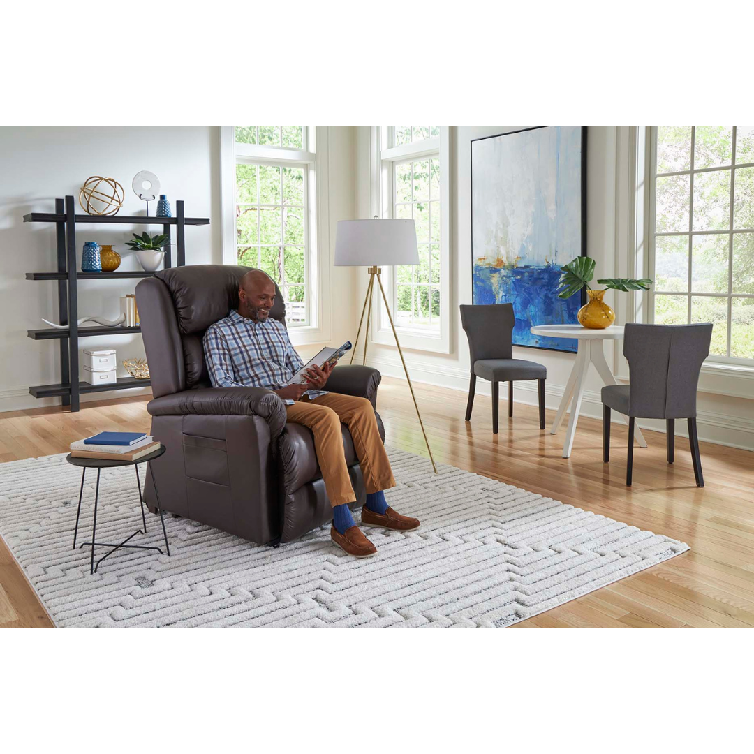 A person relaxes in a Golden Tech PR766 Relaxer MaxiComfort Recliner With Lift Assist - Small, reading a book in a bright, modern living room with large windows. The space includes a white rug, floor lamp, small table with potted plant, painting, and decorative shelf items.