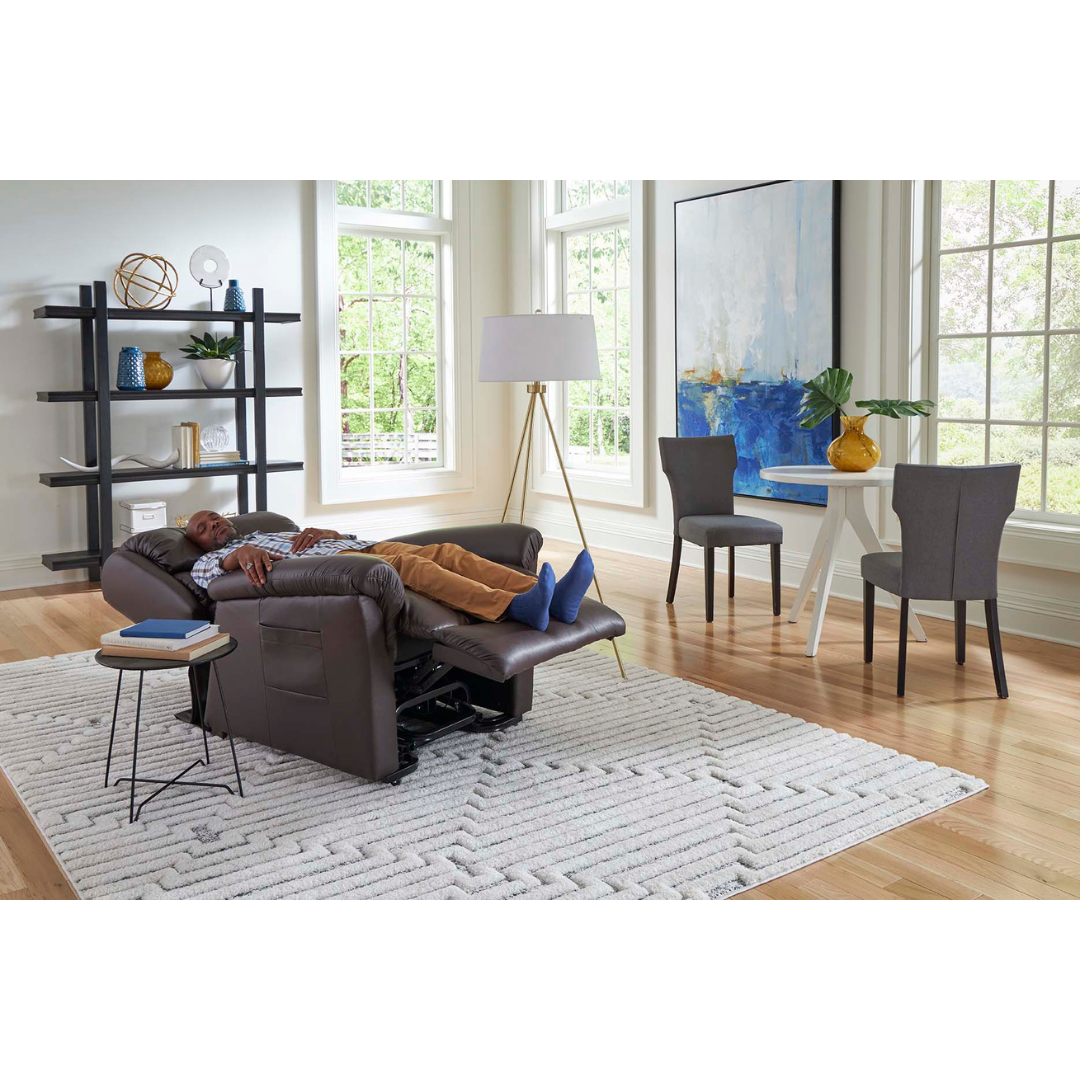 A person is peacefully sleeping on a Golden Tech PR766 Relaxer MaxiComfort Recliner with Lift Assist - Small in a bright living room with large windows. The MaxiComfort Positioning ensures ultimate comfort among the stylish rug, black bookshelf, floor lamp, painting, small round side table, and cozy dining set for two.
