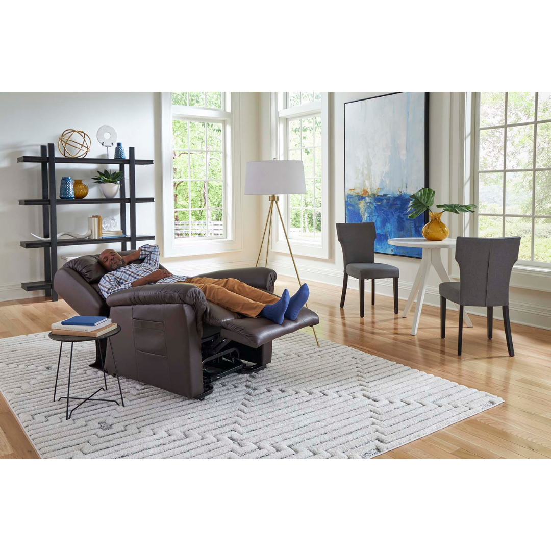 In a bright living room with large windows, a person relaxes on the Golden Tech PR766 Relaxer MaxiComfort Recliner With Lift Assist - Small. The room features a plush white rug, black shelving unit, standing lamp illuminating a round side table and dining table with gray chairs, and an abstract painting on the wall.