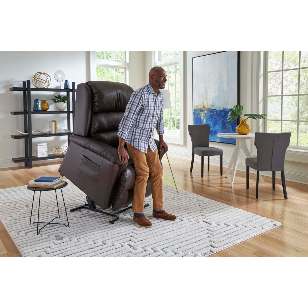 A person is standing up from a Golden Tech PR766 Relaxer MaxiComfort Recliner With Lift Assist - Small, which is in a raised position. The bright living room has a large window, white rug, small table with a plant, and a decorated bookshelf.