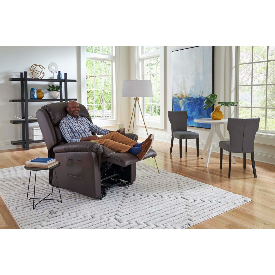 A person lounges in a Golden Tech PR766 Relaxer MaxiComfort Recliner with Lift Assist - Small in a modern living room. Large windows, a geometric rug, and a round table with chairs fill the space. A notable painting is nearby, while the programmable AutoDrive Hand Control offers ultimate comfort.