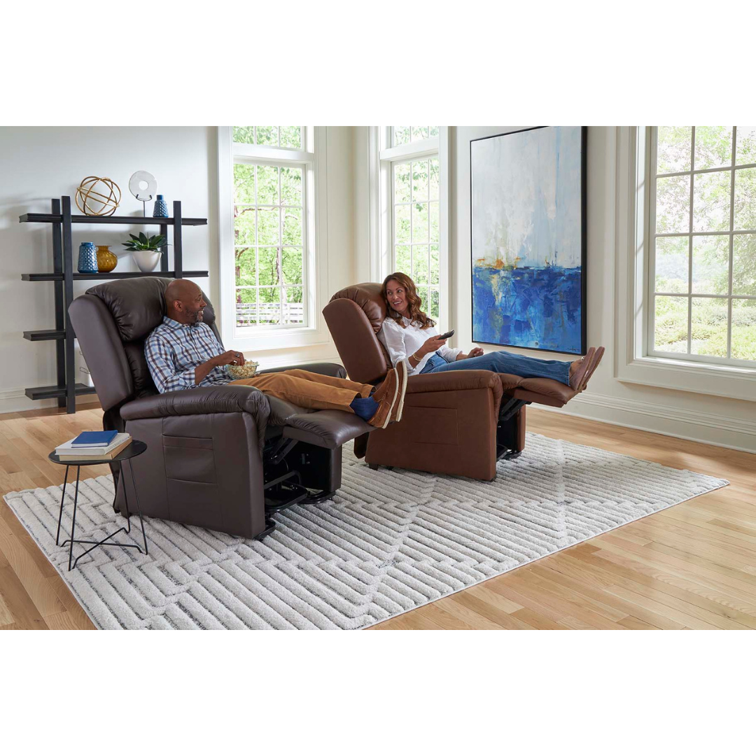A man and woman relax on Golden Tech PR766 Relaxer MaxiComfort Recliners With Lift Assist in a bright living room with large windows. They read and chat, surrounded by wooden floors, a gray textured rug, a side table, decor shelves, and a large abstract painting.