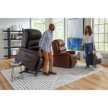 A man in a plaid shirt adjusts the Golden Tech PR766 Relaxer MaxiComfort Recliner With Lift Assist - Small near large windows. A smiling woman carries shopping bags. The cozy room features a white rug, bookshelf, and abstract artwork. The recliner boasts MaxiComfort Positioning and Programmable AutoDrive Control.
