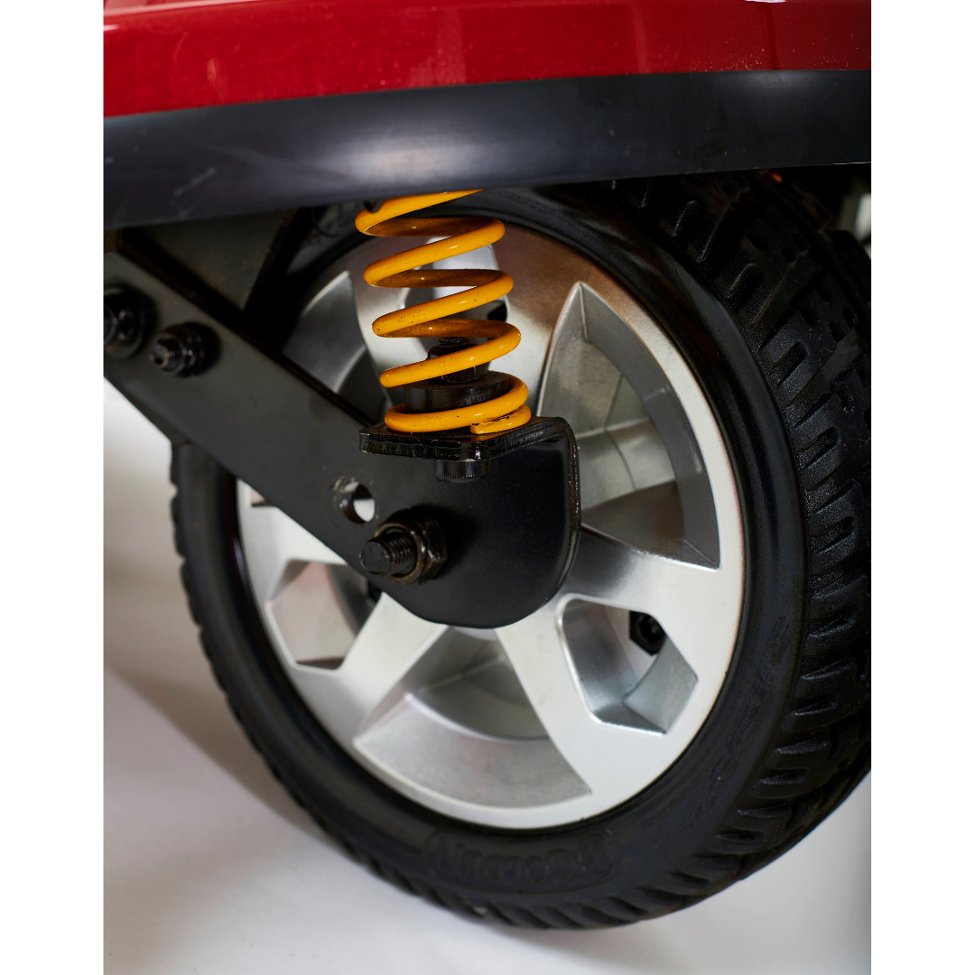 Close-up of a wheel with a silver alloy rim on the Golden Tech GC340 Companion Full-Size Bariatric Mobility Scooter, showing a yellow coil spring suspension and a black, textured tire. The vehicle body above the wheel appears partially red.