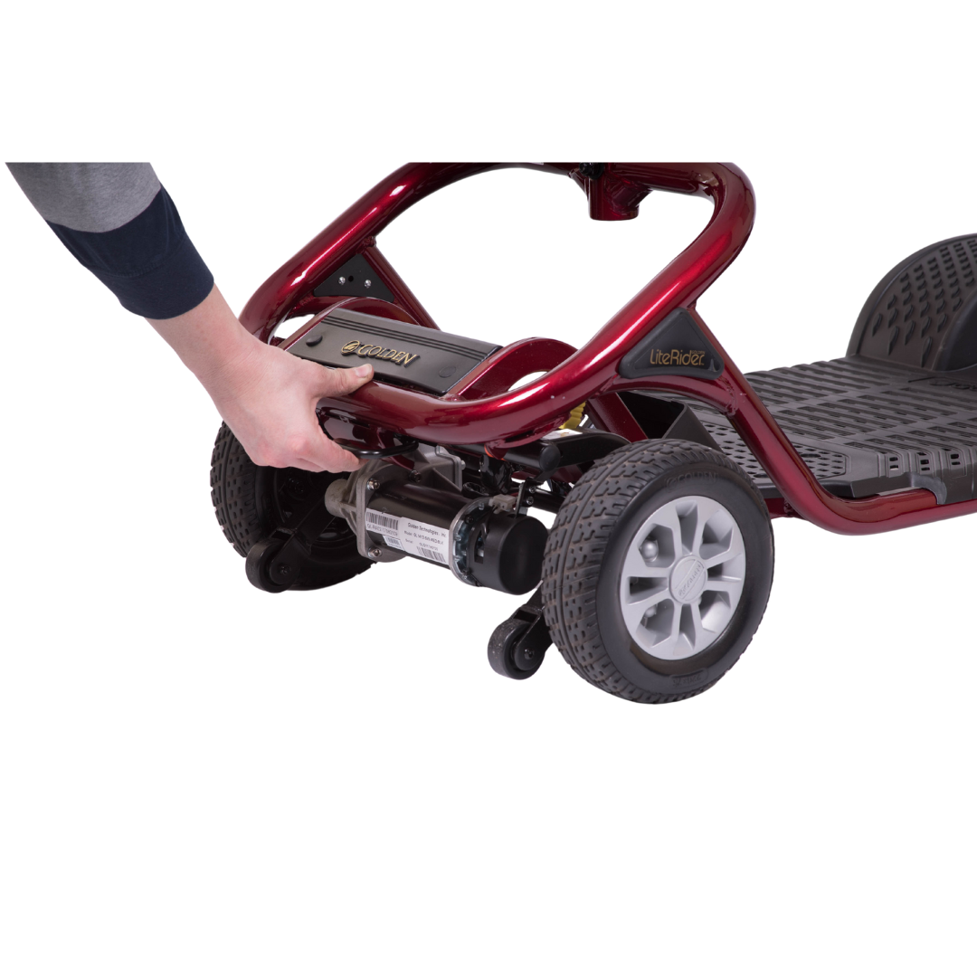 A person adjusts the back of a red Golden Tech GL111 LiteRider 3-Wheel Mobility Scooter. The hand is by the rear wheel and mechanism. This scooter, with large grey wheels and textured footrest, offers seamless wireless disassembly for easy transport.