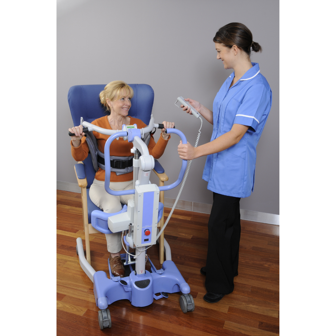 A nurse in blue helps an older woman use the Joerns Hoyer Journey® Sit-To-Stand Folding Electric Patient Lift with adjustable sling, operating the controls to improve efficiency in a room with wood floors and a gray wall.