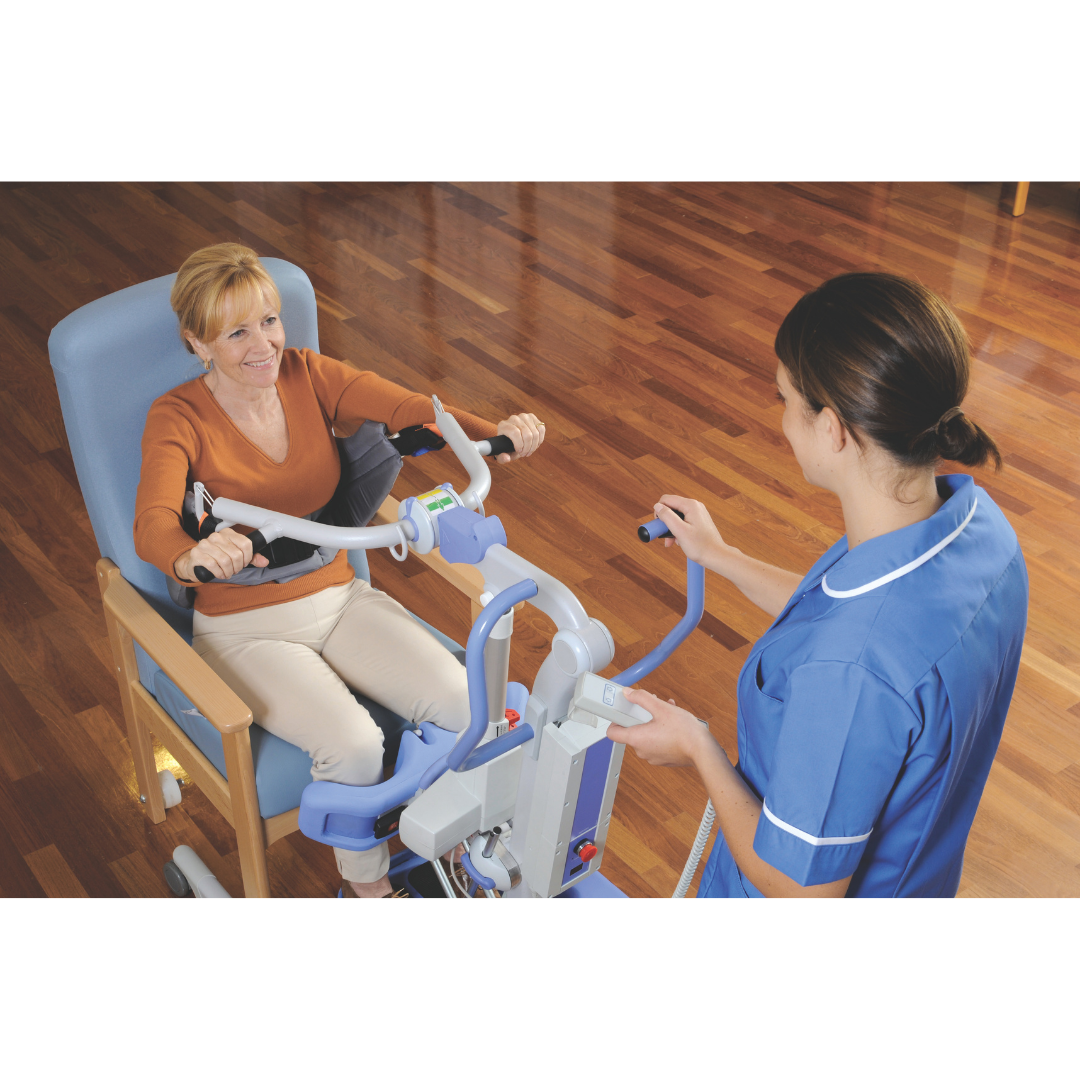 A nurse in blue scrubs uses the Joerns Hoyer Journey® Sit-To-Stand Folding Electric Patient Lift with an adjustable sling to efficiently assist a smiling, seated woman in a room with a wooden floor.