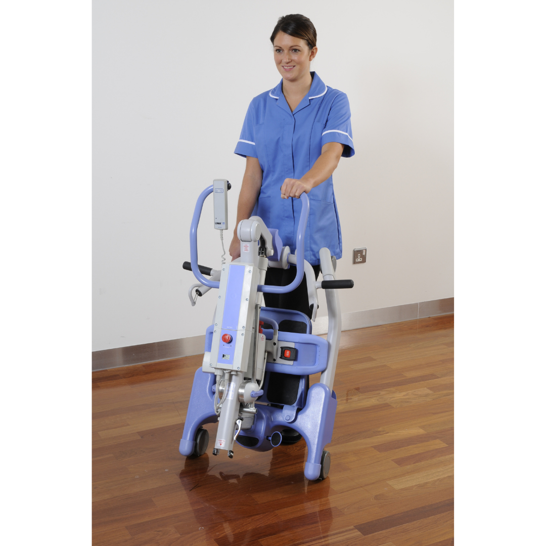 A woman in a blue nurse uniform smiles on a wooden floor, holding the Joerns Hoyer Journey® Sit-To-Stand Folding Electric Patient Lift with wheels, padded armrests, and an adjustable sling for improved caregiver efficiency.