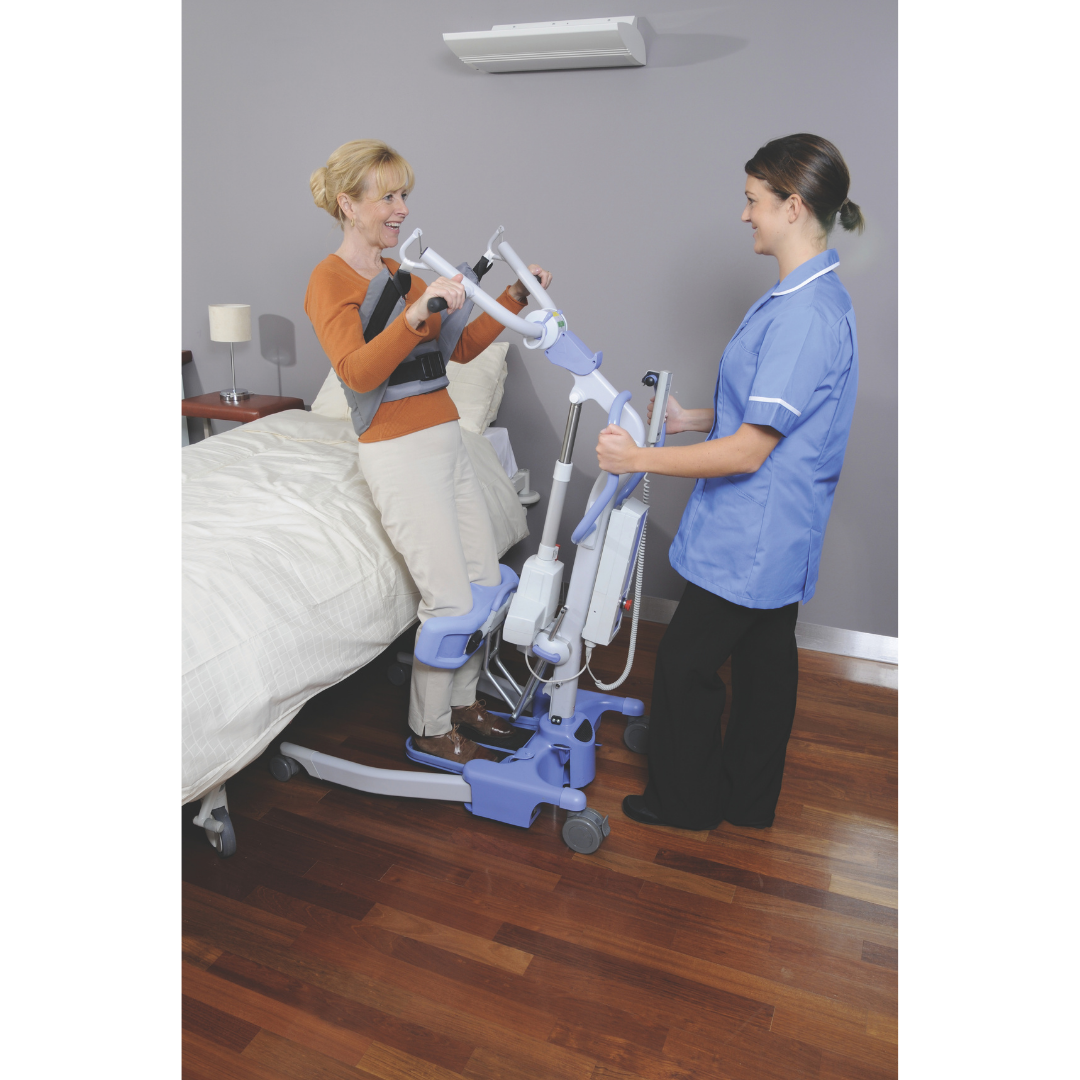 A healthcare worker assists a smiling woman using the Joerns Hoyer Journey® Sit-To-Stand Folding Electric Patient Lift with an adjustable sling attachment by the bed, supporting her as she stands and enhancing caregiver efficiency.