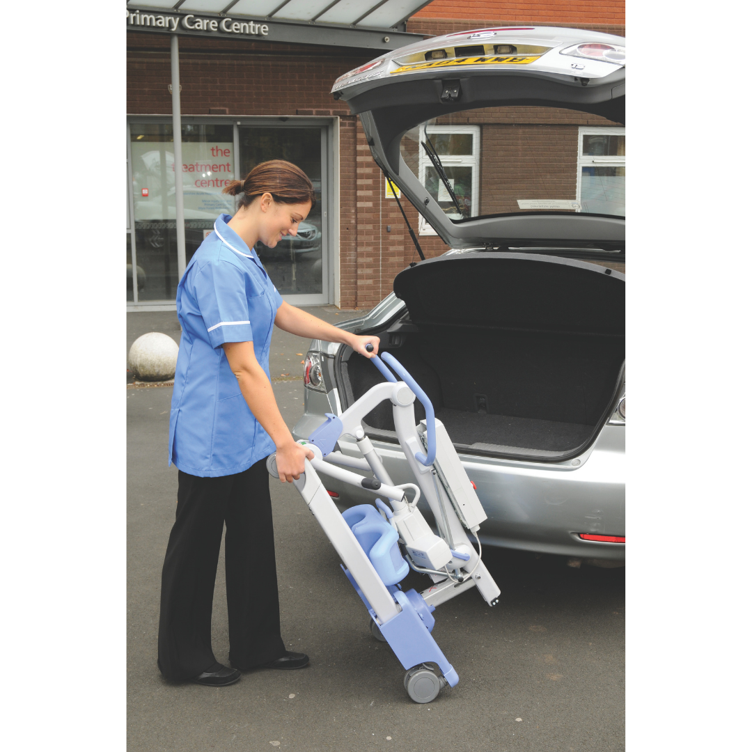 A woman in a blue uniform loads the Joerns Hoyer Journey® Sit-To-Stand Folding Electric Patient Lift into a silver cars open trunk outside a primary care center, improving caregiver efficiency.