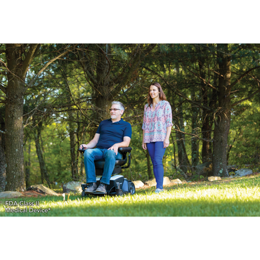 A man smiles in a Pride Mobility Go Chair® Portable Powerchair with a compact turning radius on grass, while a woman stands beside him. They are outdoors among tall trees on a sunny day. FDA Class II Medical Device appears in the lower left corner.