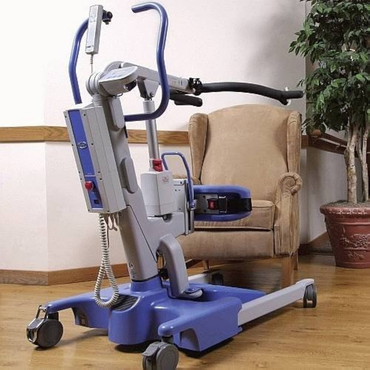 The Joerns Hoyer Elevate® Sit-To-Stand Electric Patient Lift with Smart Technology, in blue and gray with wheels and handles, is shown by a beige armchair on a wood floor, promoting caregiver safety. A potted plant appears in the background.