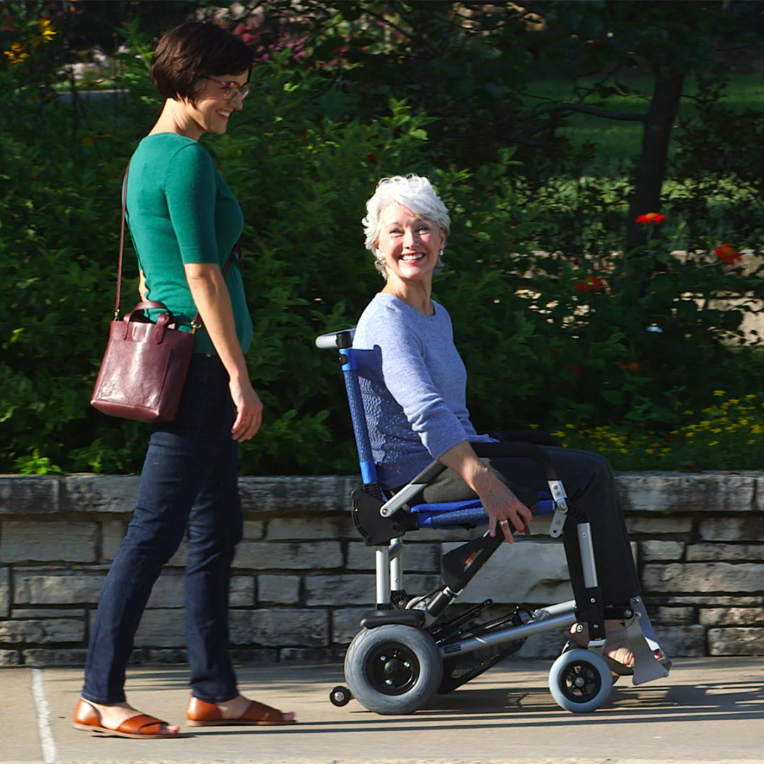 A woman with short gray hair is seated in a Journey Zinger® Folding Powerchair (FAA Approved, 47 lbs), smiling as she moves along a path. Beside her walks another woman with short dark hair, also smiling. They are outdoors, surrounded by lush greenery and flowers.