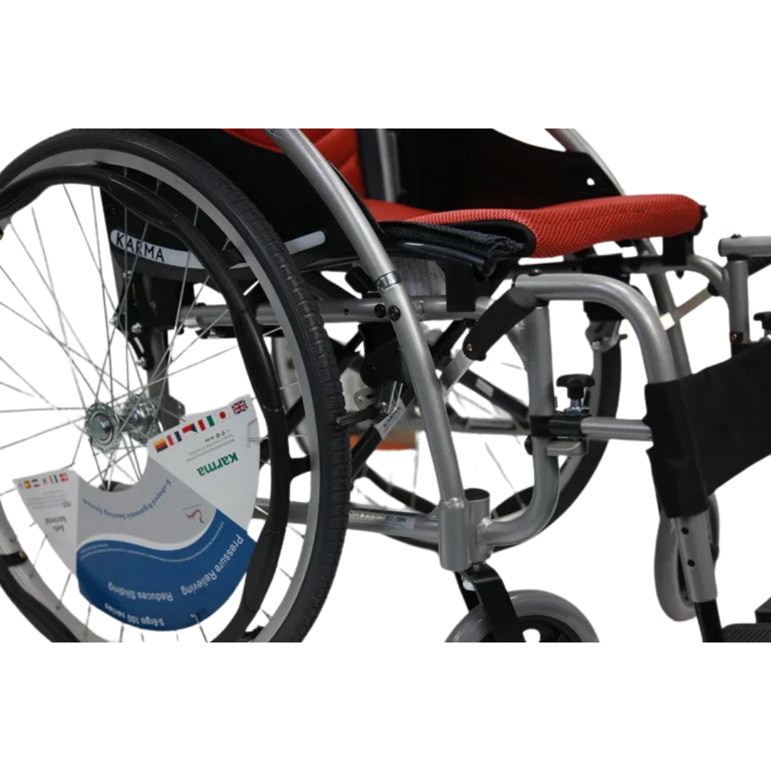 Close-up of the Karman S-Ergo 125, an ergonomic lightweight wheelchair with a metallic frame and black cushion. It has large rear wheels, smaller front wheels, a visible footrest, and flip-back arms. A label is attached to one wheel.