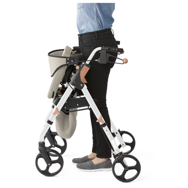A person in a denim shirt and black pants uses the Medline Empower Rollator with Microban, offering mobility assistance with large wheels and a fabric bag. Its sleek white frame with black and wooden accents provides both style and support.