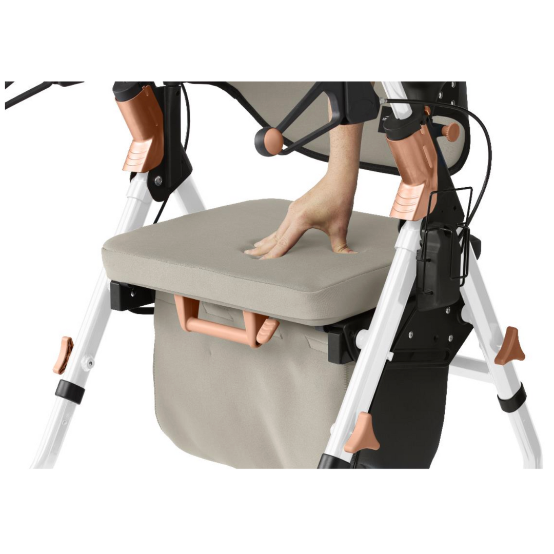 The hand pressing down on the memory foam seat of the Medline Empower Rollator with Microban illustrates its softness, complemented by a beige fabric, black and white frame, copper accents, and a convenient storage pocket hanging beneath for added utility.