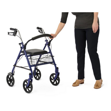 A person stands beside a Medline Steel Rollator with 8 wheels and basket. They are in a dark top and black pants, with one hand on the handle. The blue walker features hand brakes, a padded seat, and backrest for added comfort.
