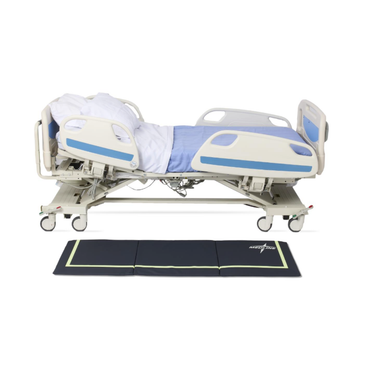 A hospital bed with side rails is in a semi-reclined position, covered with a blue and white blanket. Next to it on the floor is a Medline Trifold Beveled Edge Fall Mat with Glow featuring green edges.