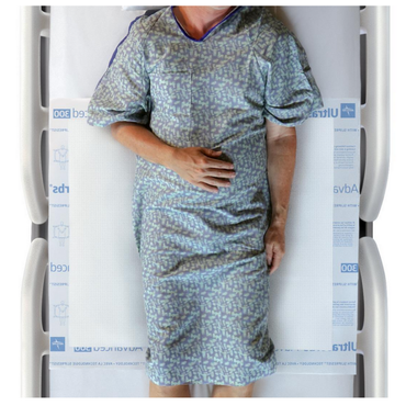 A person in a patterned hospital gown lies on their back in bed with side rails, one hand on their abdomen. The bed features a Medline Ultrasorbs Advanced Premium Underpad (30 x 36) to support effective incontinence management.