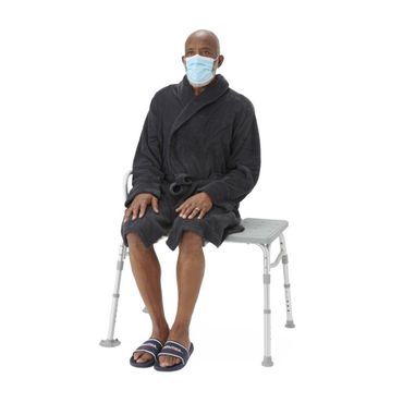 An older man wearing a face mask, black bathrobe, and slippers sits on a Medline Bariatric Transfer Bath Bench with Back—a gray shower chair with a blow-molded seat—against a white background.