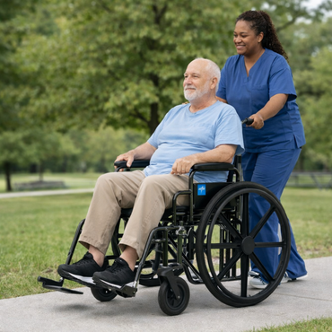 A smiling caregiver in blue scrubs pushes an elderly man in a Medline Guardian K4 Wheelchair with Desk-Length Arms along a sunny park pathway, with green trees and grass in the background.