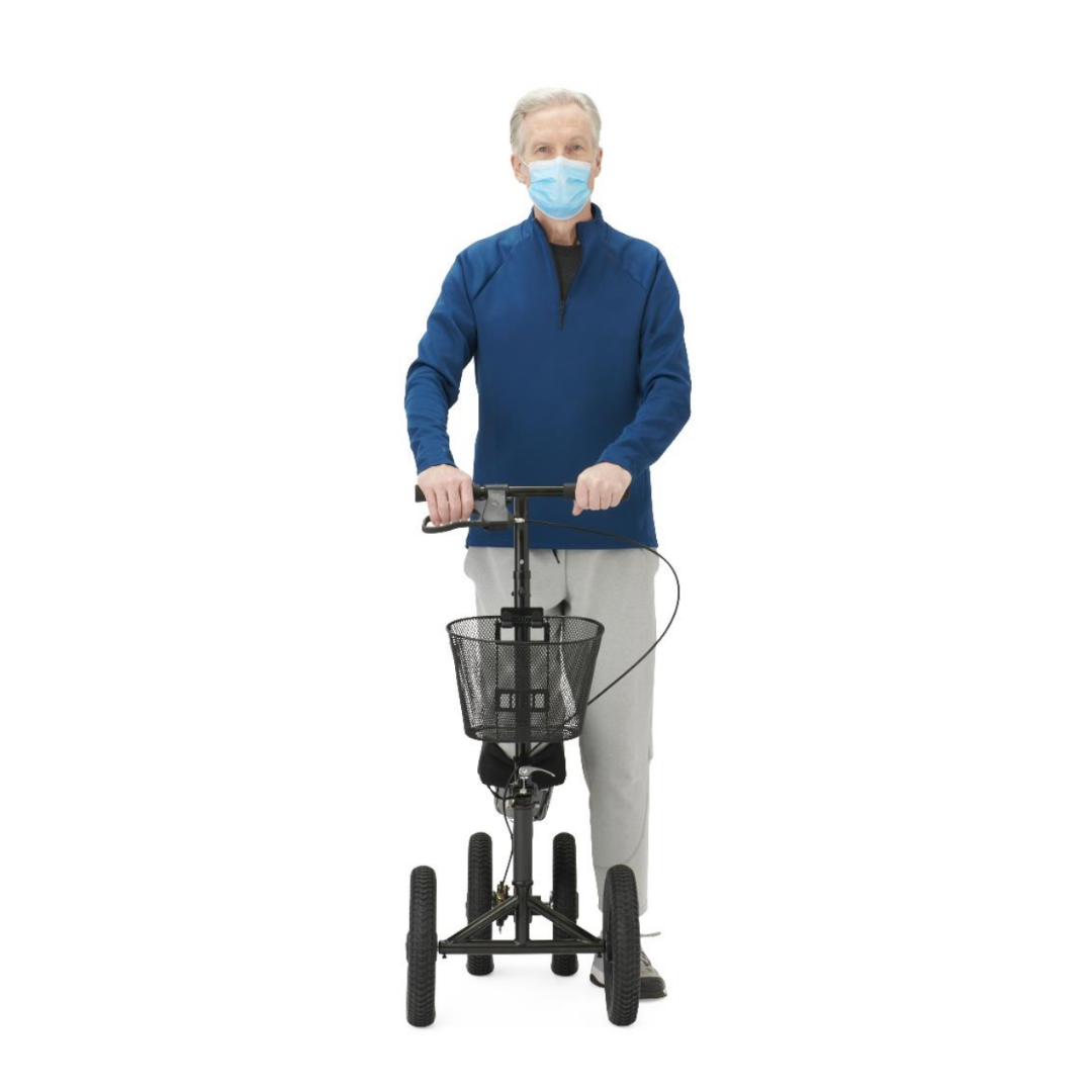 An older man in a blue jacket, gray pants, and a blue face mask stands using the Medline All-Terrain Portable Knee Walker with large wheels and a basket, set against a plain white background.