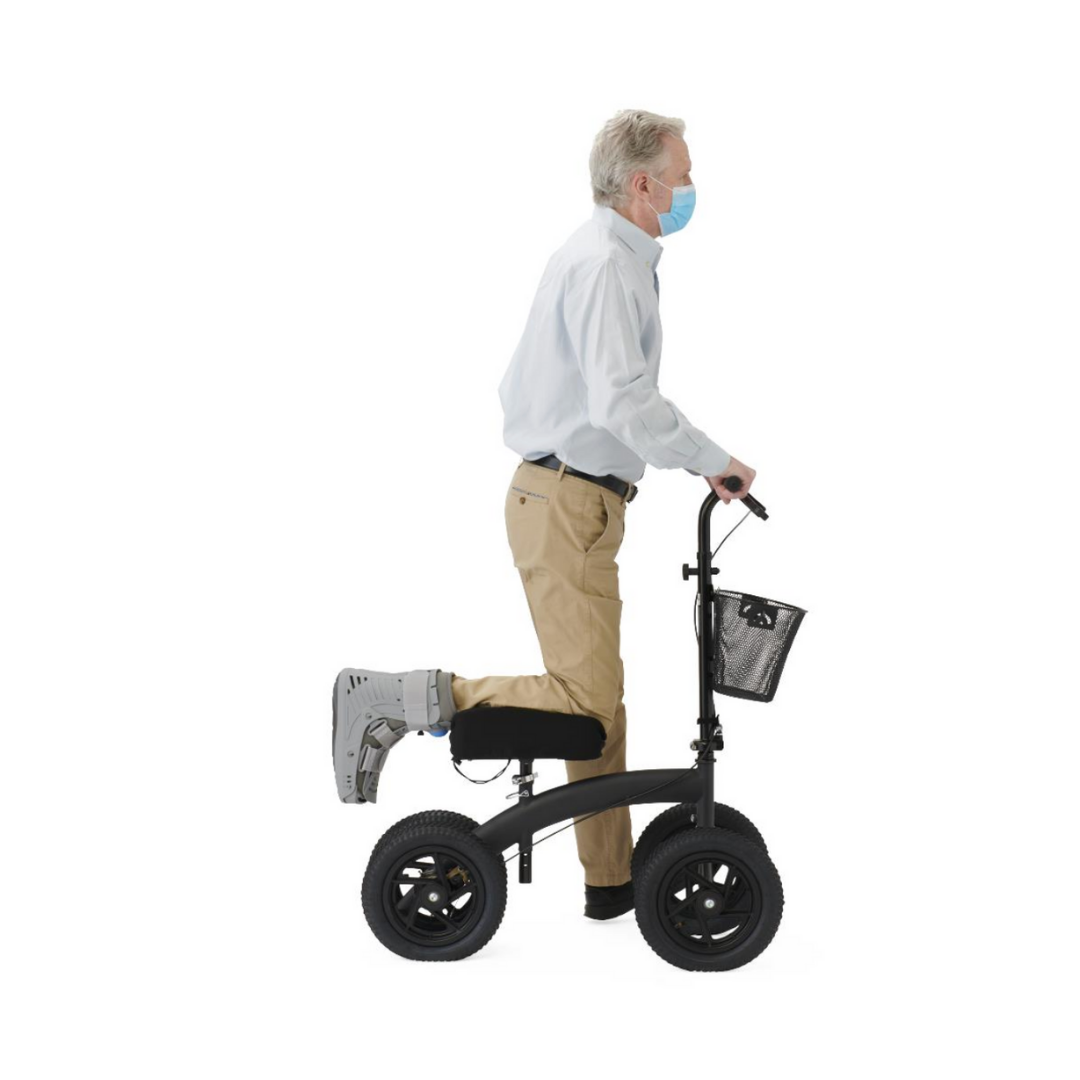 An older man in a medical mask and right walking boot uses the Medline All-Terrain Portable Knee Walker with a basket, resting his injured leg on it while standing on his left leg.