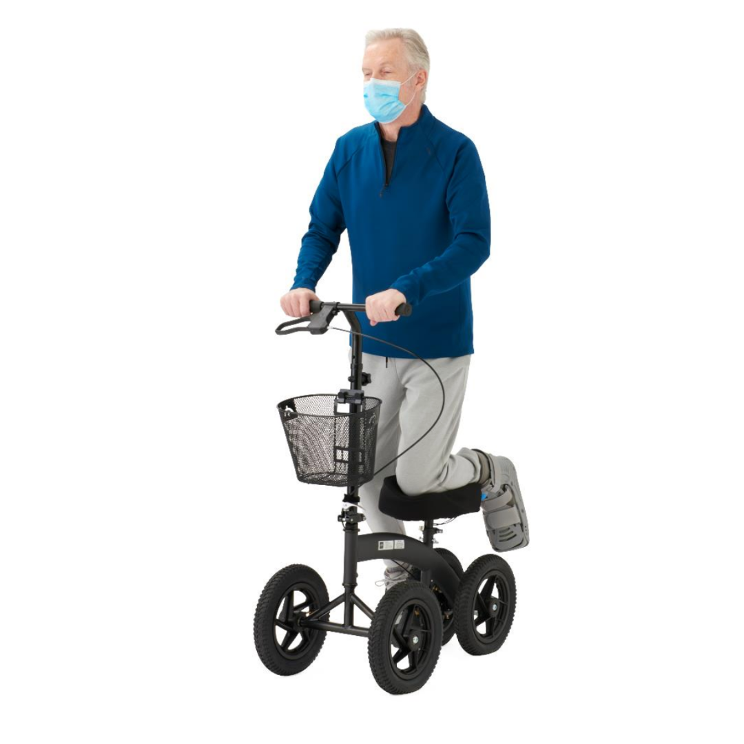 An older man in a blue zip-up jacket and light gray pants uses the Medline All-Terrain Portable Knee Walker, resting his right leg—secured in a medical boot—on its padded platform. He wears a face mask and utilizes the attached basket.