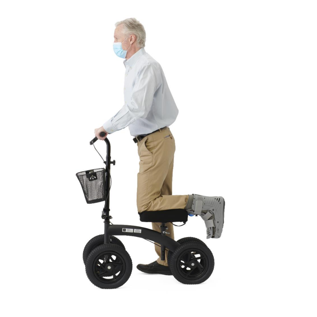 An older man in a mask and medical boot uses the Medline All-Terrain Portable Knee Walker, resting his injured leg on its padded platform with rugged wheels, against a plain white background.