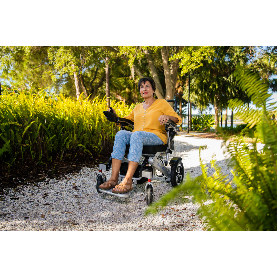 A woman in a yellow blouse and patterned pants smiles while using a Miracle Mobility Platinum 8000 Bariatric Portable Powerchair on a gravel path surrounded by green plants and trees in a sunny outdoor setting.