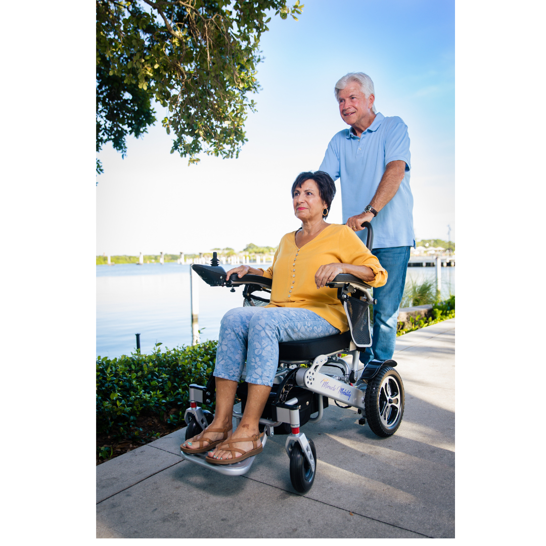 An older woman in a yellow cardigan sits in a Miracle Mobility Platinum 8000 Portable Powerchair - Open Box by a lakeside path, while an older man stands behind her smiling, holding the handles. Trees and water appear in the background.