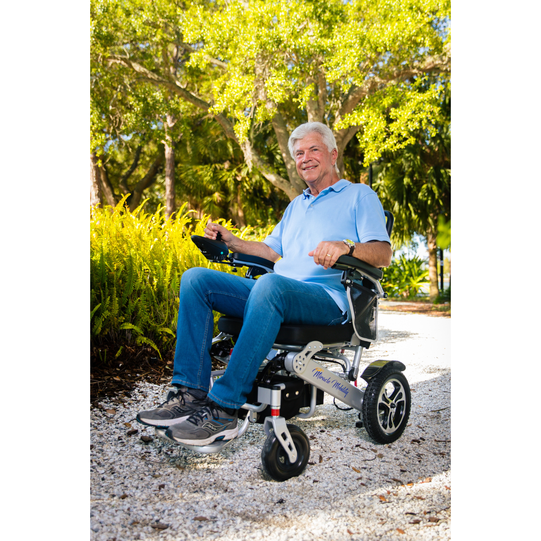 An older man with gray hair smiles while sitting in a Miracle Mobility Platinum 8000 Bariatric Portable Powerchair on a sunny gravel path, surrounded by green trees and plants, wearing a light blue polo shirt and jeans.