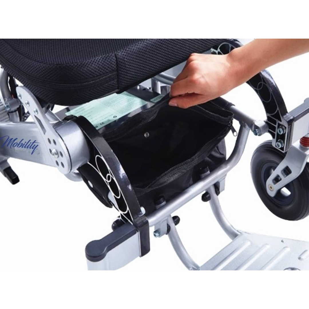 A close-up shows a hand opening a zippered storage pouch under the seat of the Miracle Mobility Platinum 8000 Portable Powerchair - Open Box. The powerchair’s frame and wheels are also visible, highlighting its travel-friendly design.