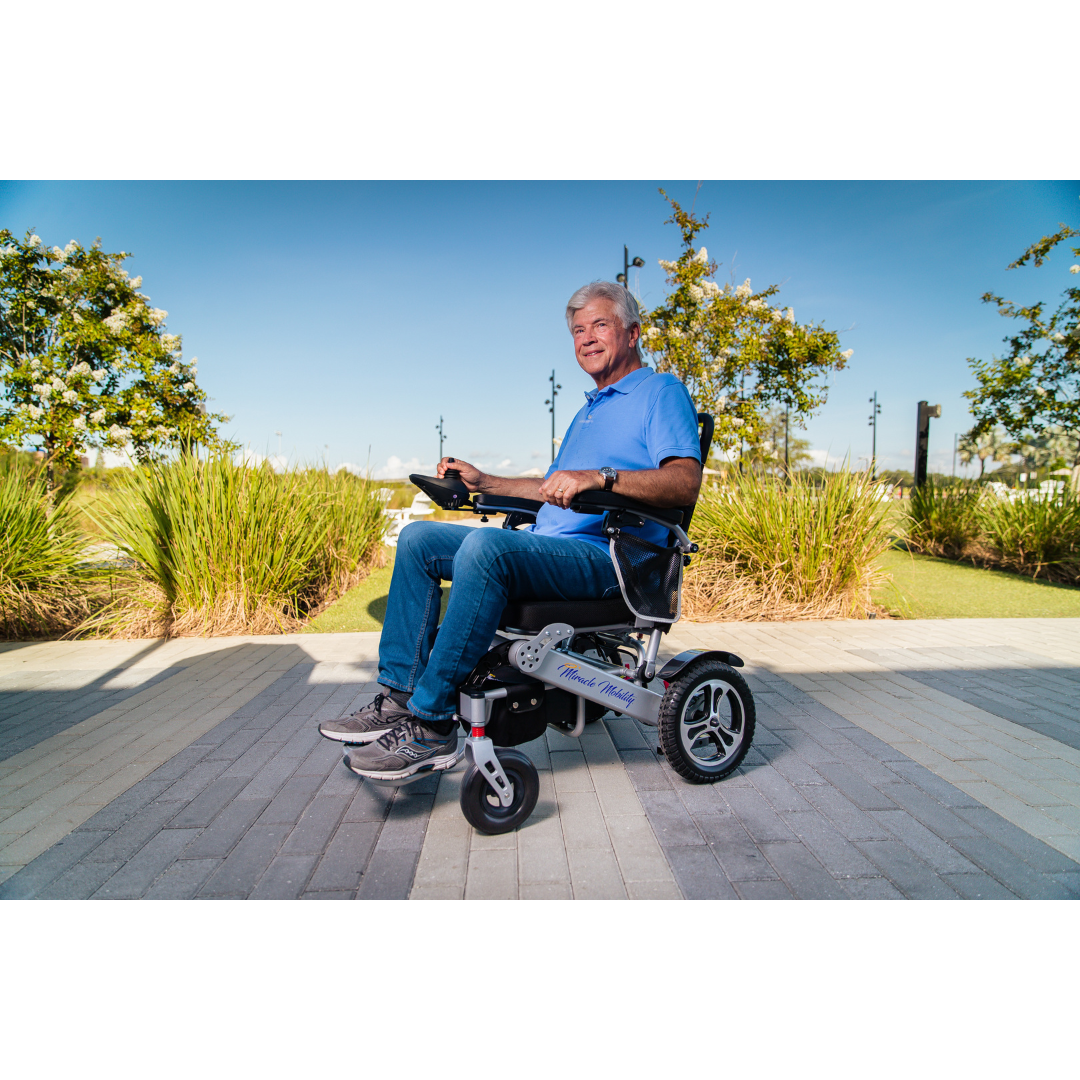 An older man in a blue shirt and jeans sits in his Miracle Mobility Platinum 8000 Bariatric Portable Powerchair on a sunny path bordered by grass and trees, smiling at the camera while holding the powerchair's controls.