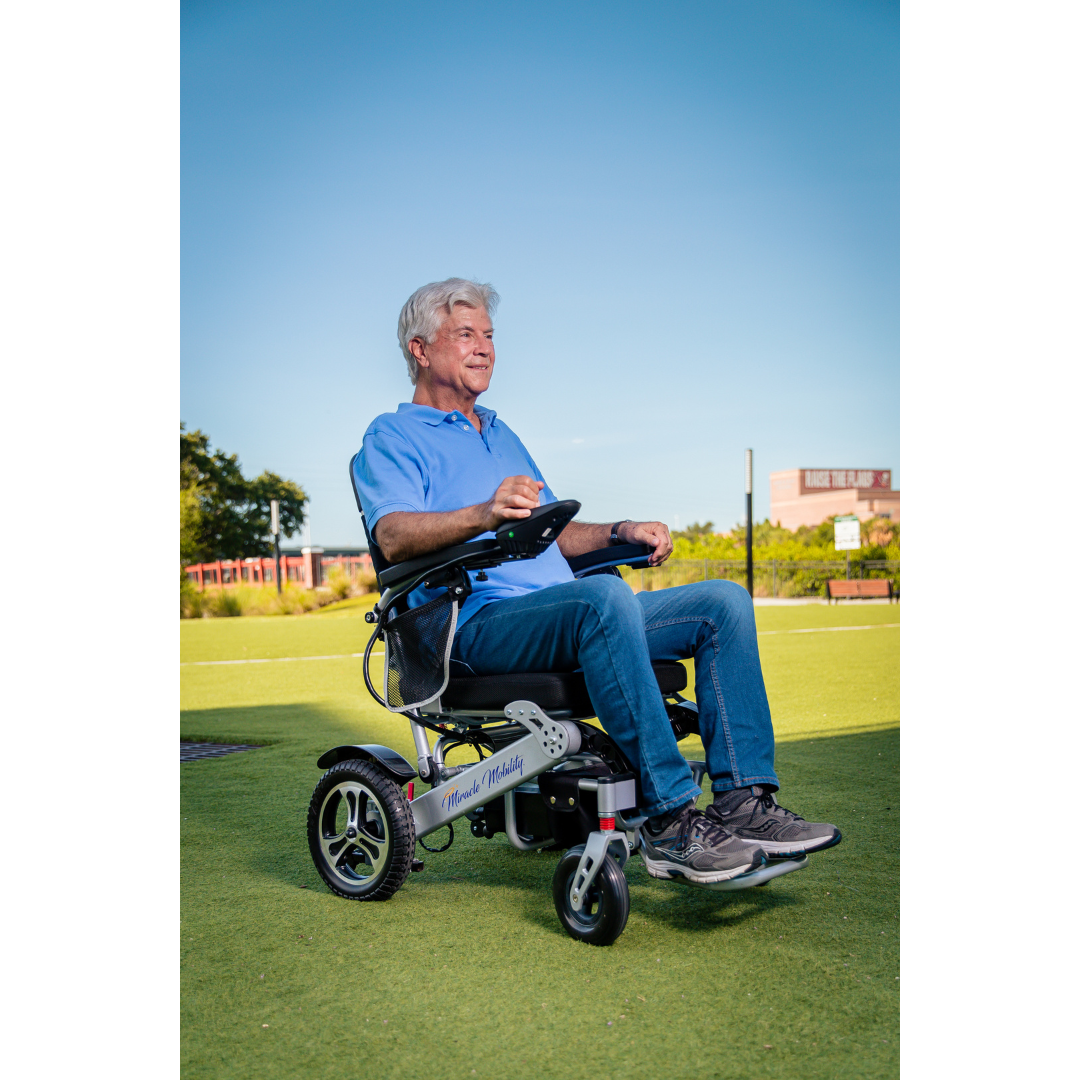 An older man with gray hair smiles while seated in a Miracle Mobility Platinum 8000 Bariatric Portable Powerchair on grass outdoors under a clear blue sky.